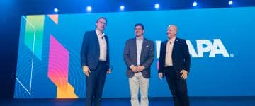 Scott O'Neil, Michael Shelton, and Jakob Wahl stand on stage together at the Leadership Breakfast, where the Best Exhibit winners were acknowledged